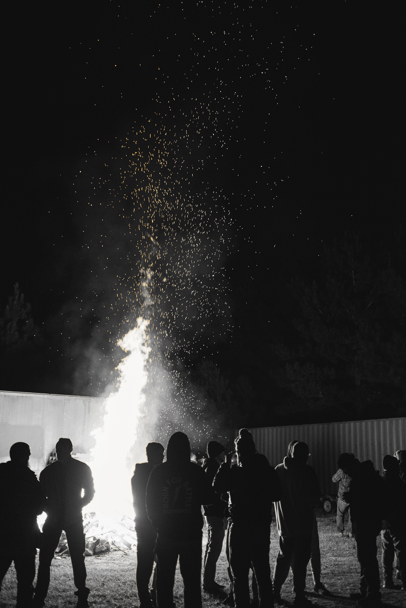 Men standing around a roaring bonfire at night during Offsite, golden sparks rising into the black sky, one man wearing a Born for Battle Brotherhood hoodie.