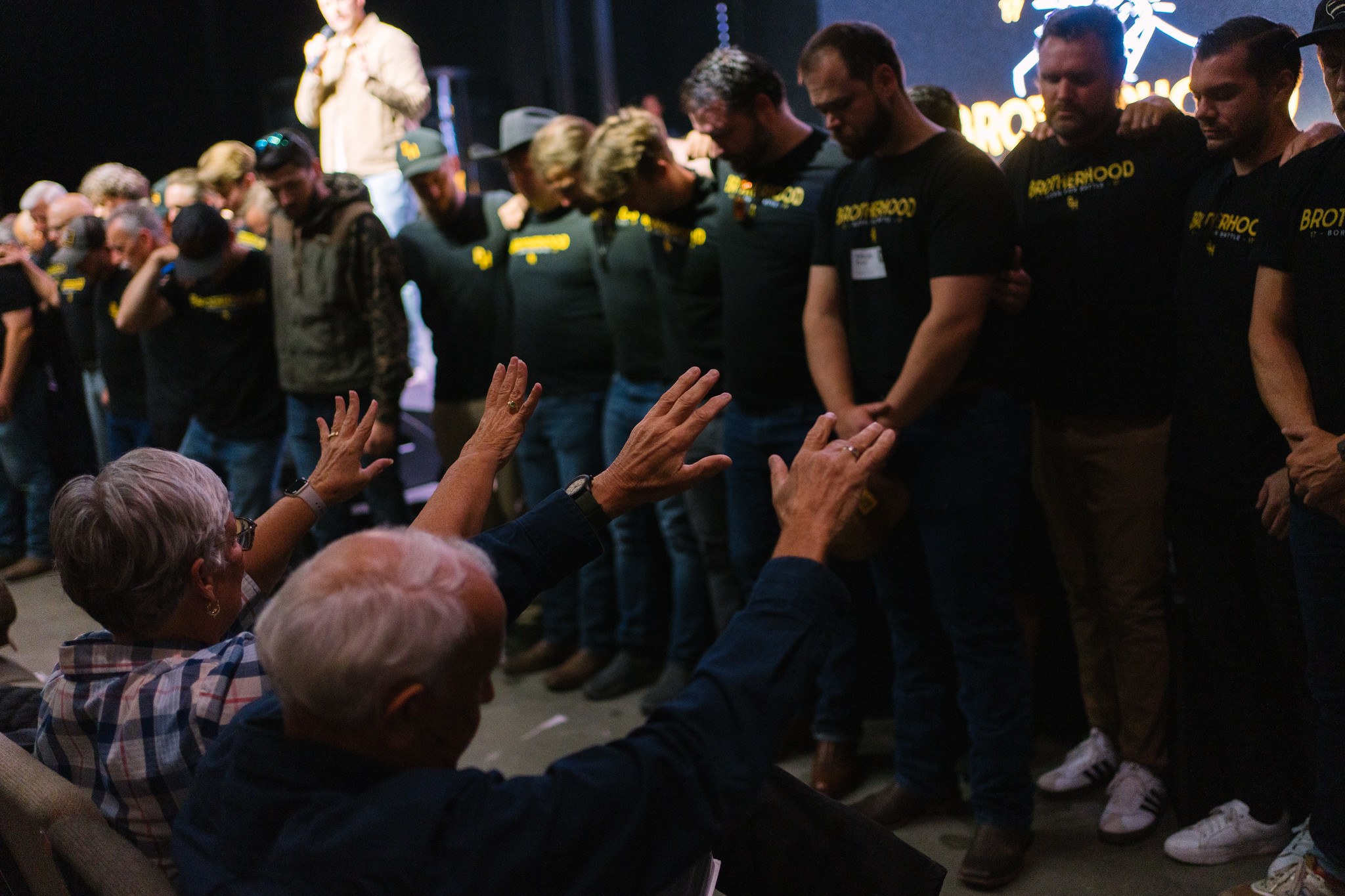 Men in Brotherhood shirts lined up with heads bowed as an older couple in the foreground raises their hands in blessing, a stage behind them.