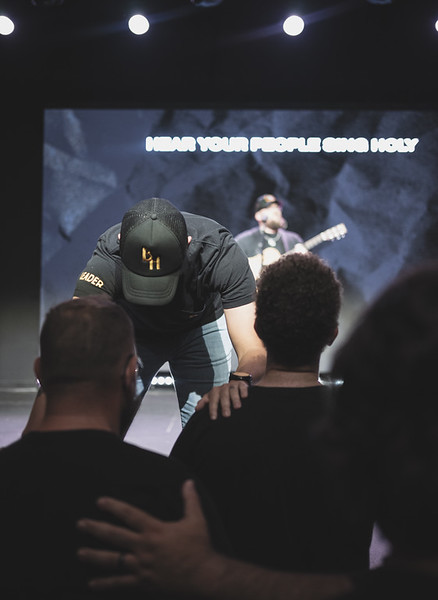 A Brotherhood leader bowed in prayer during worship, hand on his shoulder, lyrics on the screen behind