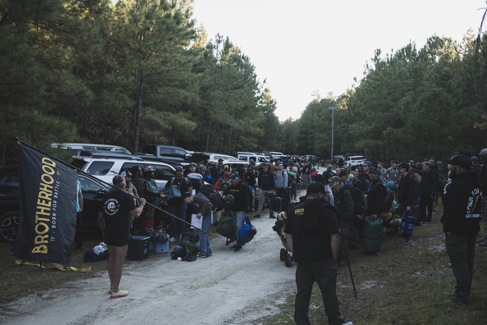 Men gathering at dawn in a sandy parking lot with a Brotherhood flag and a Born for Battle hoodie: trucks, backpacks, and pine woods.