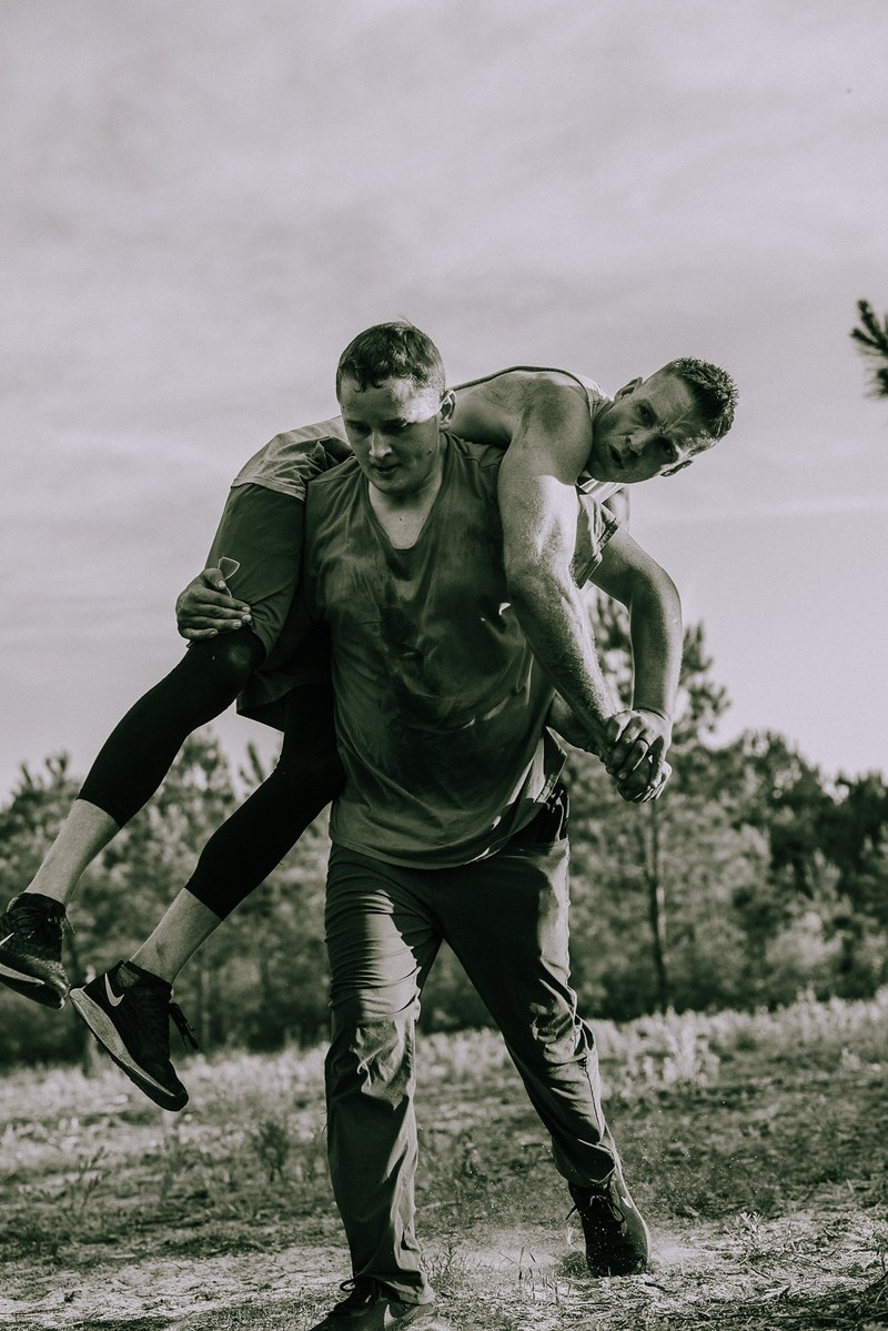 A man carrying another brother across his shoulders in a fireman's carry across a field, the carried man resting forward over the carrier's back.