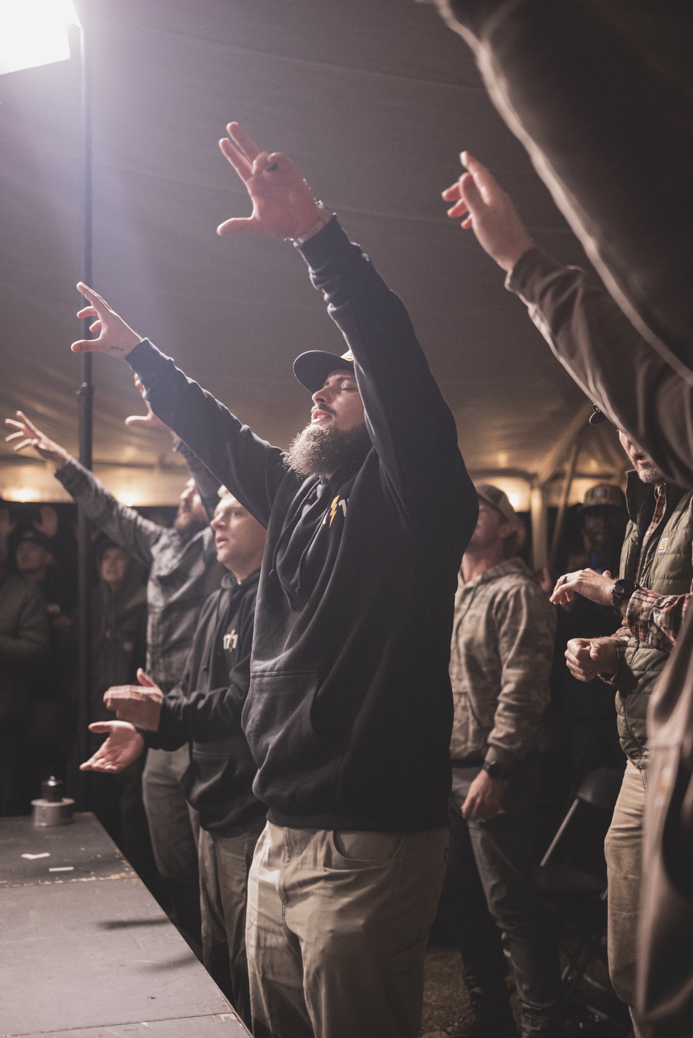 A bearded man in a black hoodie worshiping with arms raised under a tent, light flaring overhead.