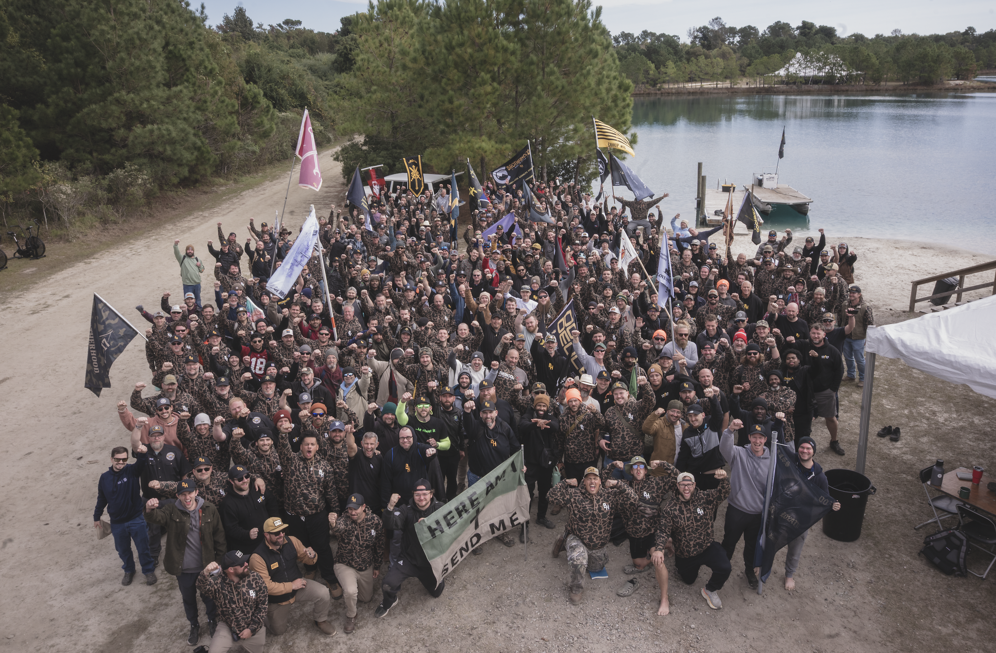 Hundreds of Brotherhood men gathered at a lakeside clearing, fists raised, holding a HERE AM I SEND ME banner: tents and flags behind them
